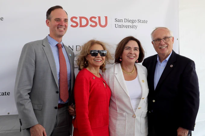 Four smiling individuals stand in front of a San Diego State University (SDSU) step-and-repeat banner. From left to right: a man in a gray suit, a woman in a red outfit with sunglasses, a woman in a white suit, and a man in a dark blazer.