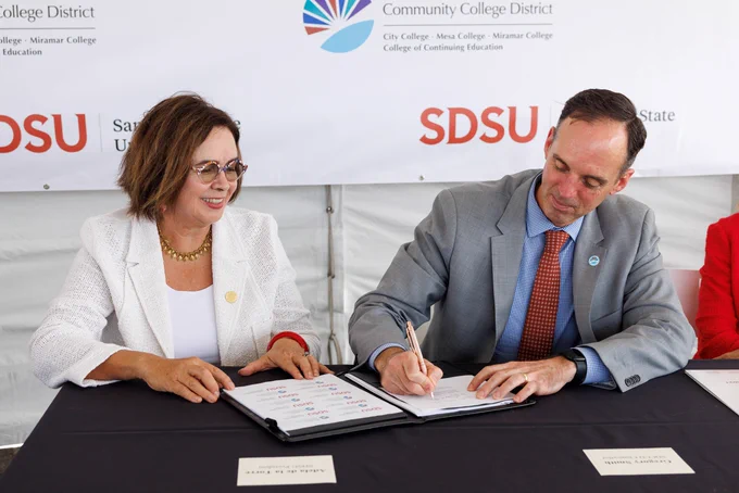 A woman in a white blazer and a man in a gray suit with a red tie sit at a table, smiling and signing documents during a formal partnership event between SDSU and the San Diego Community College District.