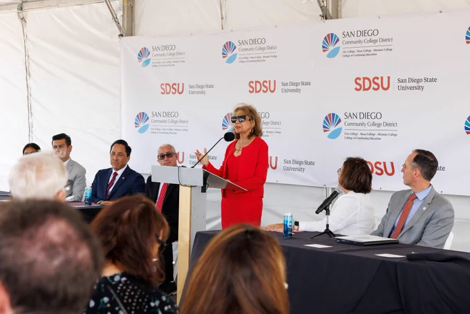 A woman in a red outfit speaks at a podium during a formal event, with a panel of seated officials behind her and a branded backdrop featuring SDSU and the San Diego Community College District logos.