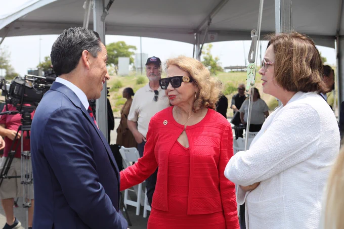 A man in a navy suit speaks with two women—one in a red outfit with sunglasses and the other in a white blazer—under an outdoor tent during a public event. People and media equipment are visible in the background.