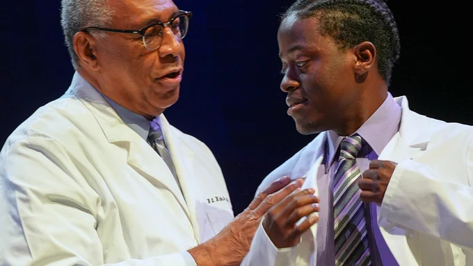 A student smiles while being helped into a white coat on the stage