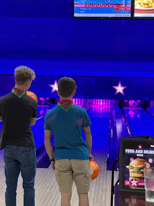 Two teenage males holding bowling balls and wearing neckerchiefs, beyond them is the bowling alley lane and ten pins ready to be knocked down.