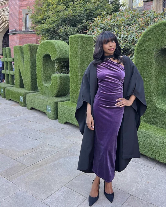 Graduate in a purple dress and black robe standing in front of a green hedge with the letters 'NCL' at National College of Ireland.