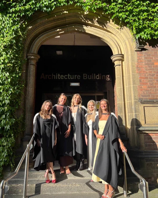 Group of five graduates smiling in front of the Architecture Building, dressed in graduation robes and formal attire.