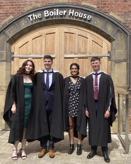 Four graduates stand in front of "The Boiler House" entrance, smiling and dressed in academic robes.