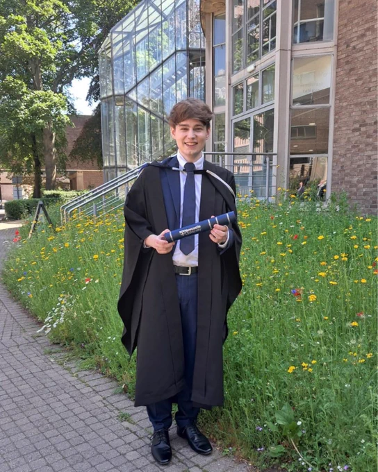 Person in graduation attire holding a diploma, standing in front of a modern building with extensive glass windows and surrounded by a wildflower garden.