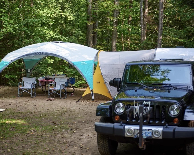A campsite set up in a forested area with a black vehicle parked at the front, a tent, and a tarp-covered area with chairs and a table.