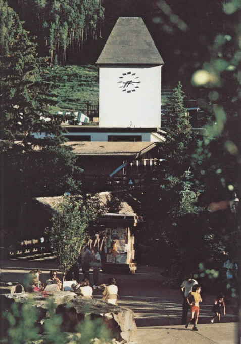 Classic view of Vail Village with covered bridge and clocktower visible, surrounded by lush green trees, people meandering.