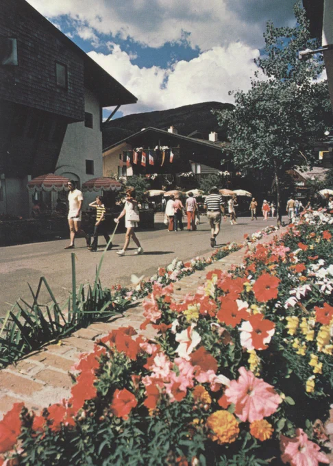 A vibrant street scene showing people strolling and mingling in a town square with blooming flowers, green plants, and umbrella-shaded tables under a clear blue sky.