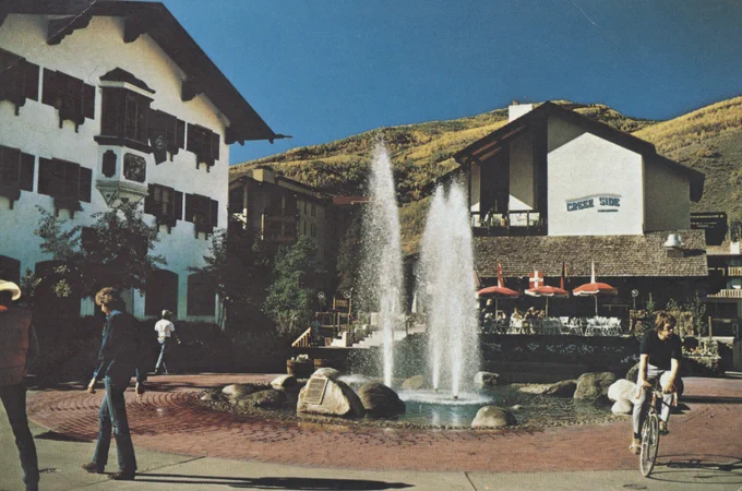 A vibrant, historic scene at Vail Village featuring a central fountain with water jets, nestled between multi-story, chalet-style buildings. People are scattered around, engaging in various activities like walking and cycling under a clear blue sky.