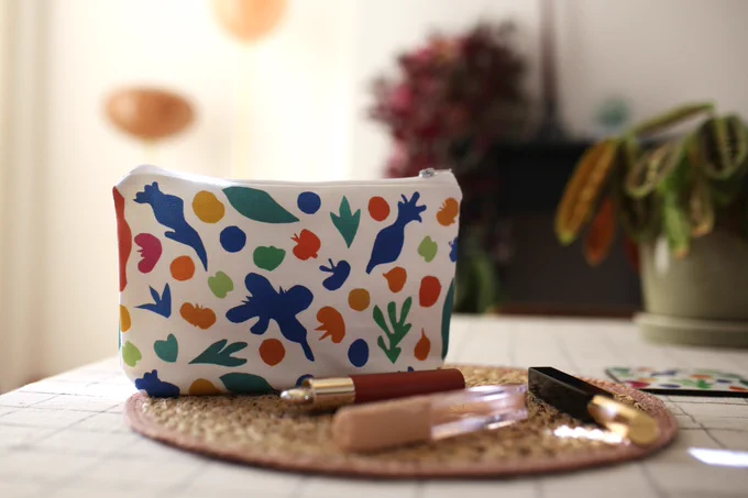 Colourful makeup bag with abstract shapes sits on a woven mat alongside makeup on a checkered tablecloth.