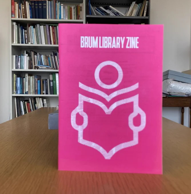 A book on a table with the front cover facing on to the camera. Title of the book at the top of the page is Brum Library Zine. Cover has a logo for reading a book on it. Behind the book is a large bookcase and a table on the left with boxes on it. 