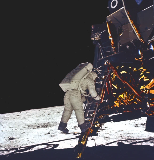 NASA astronaut Buzz Aldrin climbs down the ladder of the Apollo 11 Lunar Module “Eagle,” moments before stepping onto the Moon for the first time on July 20, 1969. He wears a white spacesuit with a rectangular life support backpack, his boots hovering just above the dusty gray lunar surface. The sky behind him is dark and vast. The lander’s metallic structure is covered in crinkled gold-colored material, reflecting the sunlight. Credit: NASA