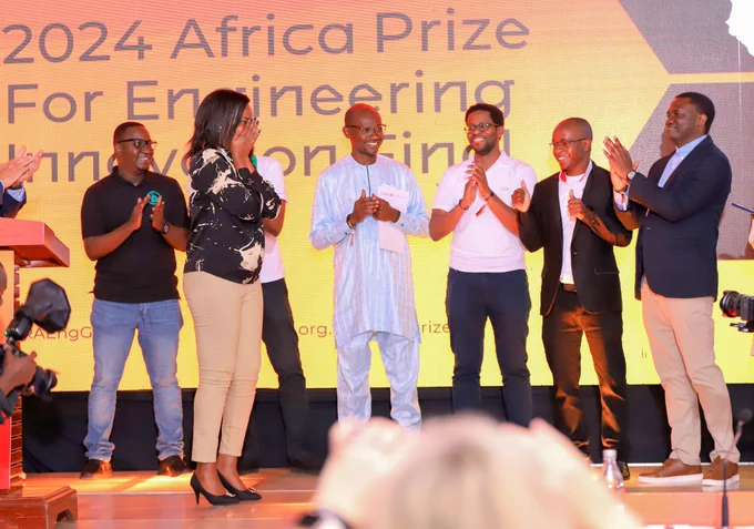 A woman joyfully holds her face in her hands while six men on stage around her applaud.