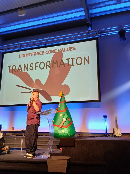 On the screen it says 
LIGHTFORCE CORE VALUES 
TRANSFORMATION 
and a picture of a butterfly. 
On the platform is Richard talking and a man in a blow up Christmas tree costume. 
