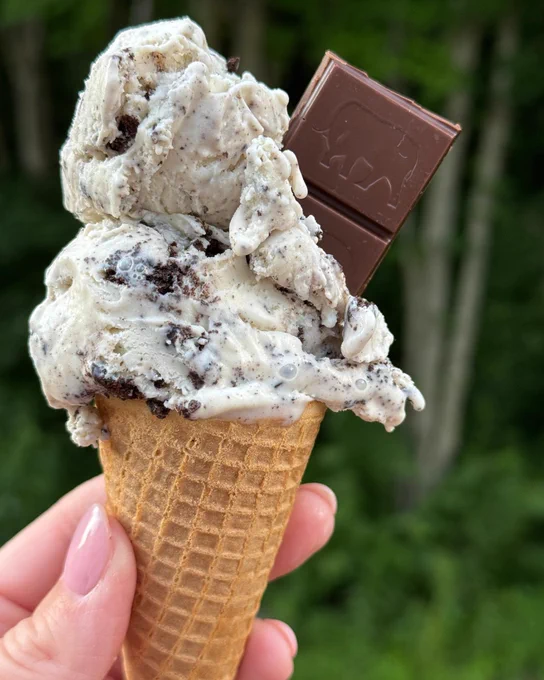 A close-up of a hand holding a cone with scoops of cookies and cream ice cream, garnished with a small piece of chocolate.