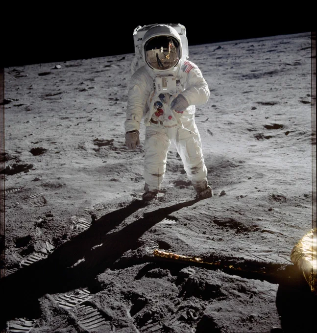 Astronaut in Apollo spacesuit standing on the Moon. Another astronaut and part of the lunar module are reflected in the gold visor of his helmet.

