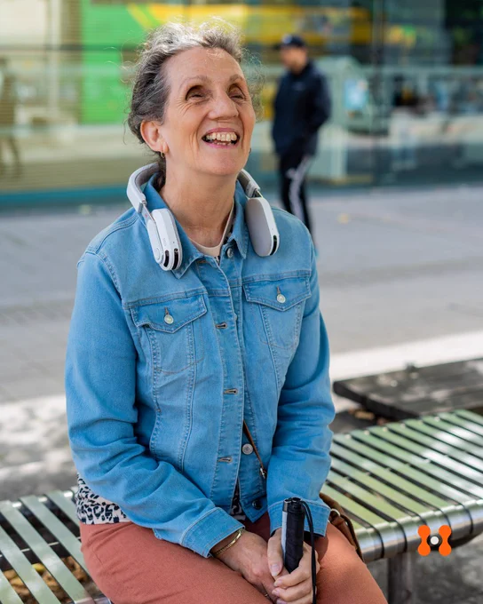 A smiling woman sits on a metal bench outdoors, wearing a light denim jacket and rust-colored pants. She has a white assistive wearable device resting around her neck and is holding a white cane, indicating she is visually impaired. Her expression is joyful as she looks upward. A blurred pedestrian and storefront are visible in the background. The orange Hope Tech logo appears in the bottom right corner.