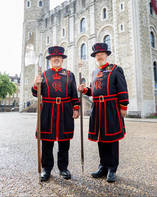 Two Yeoman Warders standing in front of the Tower of London, wearing traditional uniforms and holding ceremonial staffs.