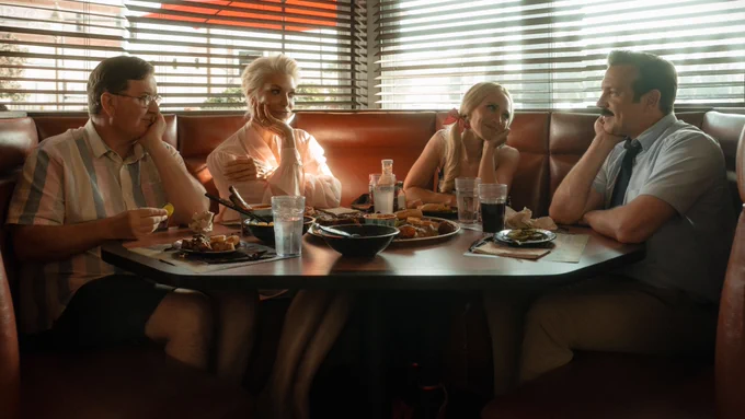Leslie Higgins, Rebecca Welton, Keeley Jones, and Ted Lasso sit side-by-side in a corner booth at a BBQ restaurant. Leslie, Rebecca, and Keeley are all looking at Ted, who is looking back at them. On the table are plates of food and glasses of water and soda.

