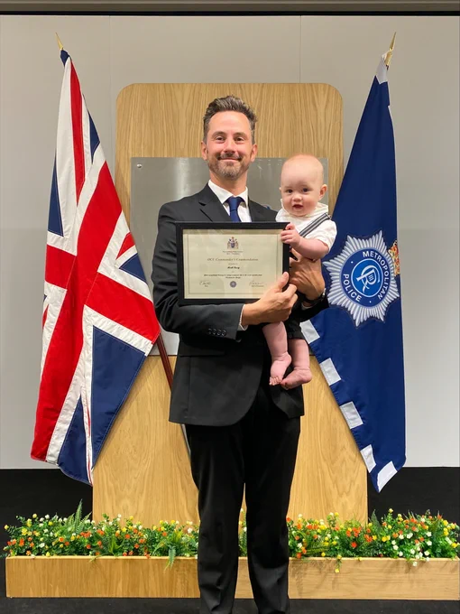 RNLI volunteer Mark holds his baby and a framed certificate, standing between the Union Jack and Met Police flags. 