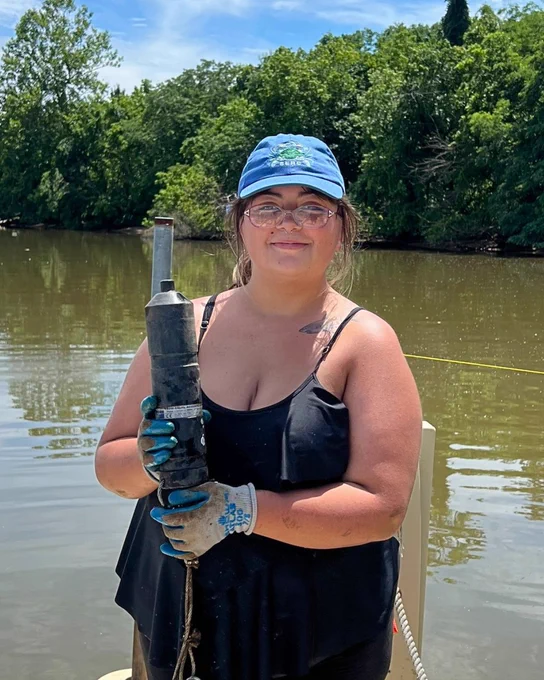 Leticia Cuevas, a young woman with dark hair and glasses, wearing a black tank top and blue hat, stands in front of a body of water holding a metal receiver.  