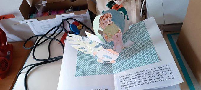 Photographie d’un livre pop-up ouvert sur une table. Deux personnages dessinés et colorés à la main surgissent en volume du livre. Au premier plan, une grande feuille découpée et un objet volant. Un texte imprimé en bas présente le personnage principal, Louna, une jeune aventurière de 18 ans. À l’arrière-plan, on aperçoit du matériel de fabrication : câbles, boîte avec bobines de filament, sac et mobilier scolaire. 