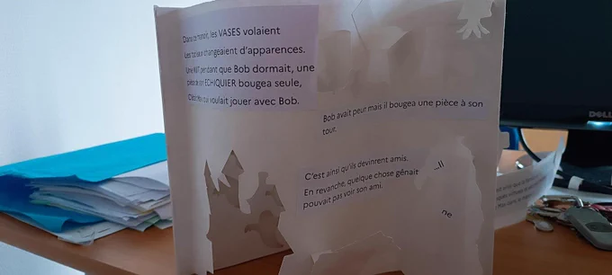 Photographie d’un livre pop-up en papier blanc, comportant des découpes en forme de château et d’autres silhouettes. Des extraits d’histoire sont collés sur les pages, racontant une scène mystérieuse où un échiquier bouge tout seul dans un manoir. En arrière-plan : un bureau avec des dossiers, des clés et un écran d’ordinateur.