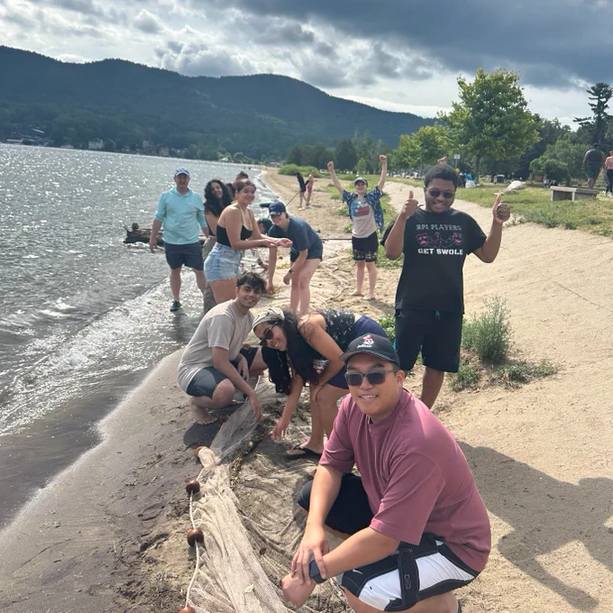 Group of students enjoying a day at Lake George with some individuals posing and smiling for the camera. The background features a calm lake and hills under a cloudy sky.