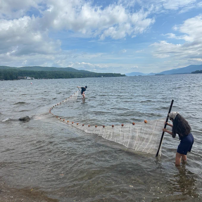 Two individuals setting a fishing net in Lake George, with mountains visible in the background on a cloudy day.