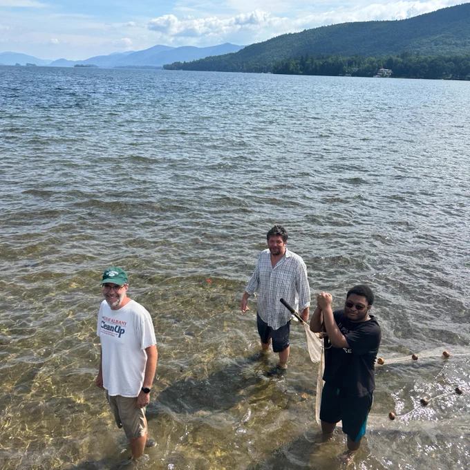 Three people standing in the shallow waters of Lake George, with mountains visible in the background. Two are smiling at the camera, and one is holding a fishing net. There is a serene landscape surrounding them.