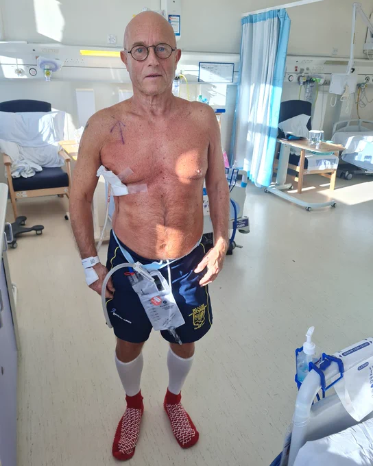 David, a white man, stands facing the camera, shirtless in a hospital room.  He has medical tubing and a drainage bag attached to his shorts.