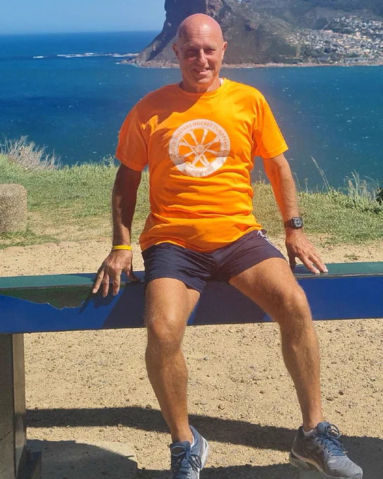 David is sitting on a bench smiling and posing for the camera overlooking a coastal landscape with rocky hills. He is wearing an orange t-shirt and navy shorts.
