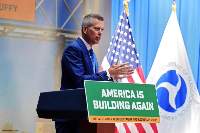 USDOT Secretary Duffy speaking at a podium with a sign that reads "America is Building Again," with U.S. and Department of Transportation flags in the background.

