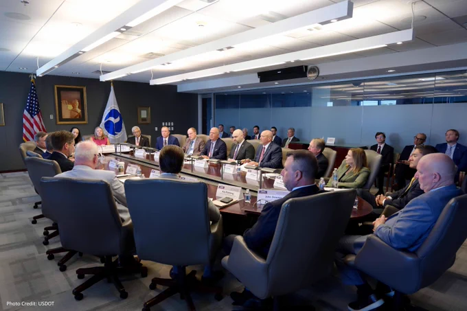 A group of individuals seated around a long rectangular conference table in a room with flags of the United States and the USDOT visible, engaged in a formal meeting.
