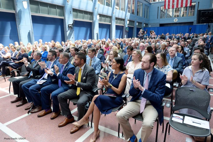 Audience at a formal event clapping, seated in a large atrium with US flag hanging from above.
