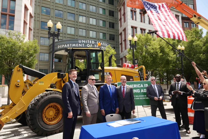 Officials at a ceremonial event in front of U.S. Department of Transportation building with a backhoe with a large American flag hanging from a crane, 
