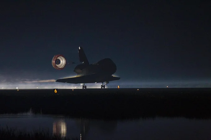 Space Shuttle landing at night with its drag parachute deployed, illuminated by runway lights.
