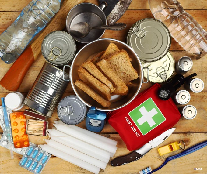 Emergency survival kit with canned food, water bottles, first aid kit, flashlight, knife, matches, and medication on a wooden surface.