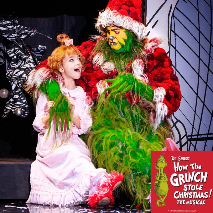A scene from "Dr. Seuss' How The Grinch Stole Christmas! The Musical" featuring the Grinch in a red suit with fluffy green fur, sitting next to a joyful child in a pink ruffled dress, both on a whimsically decorated stage.
