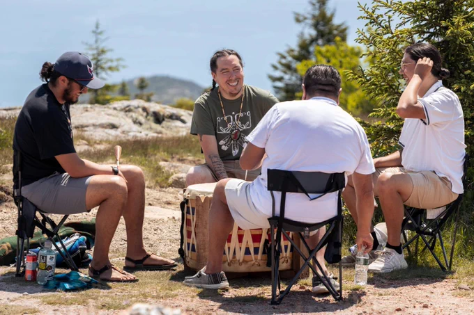 A group of four men gathered in nature, seated in a circle with a large drum at the center, surrounded by trees and rocky terrain.