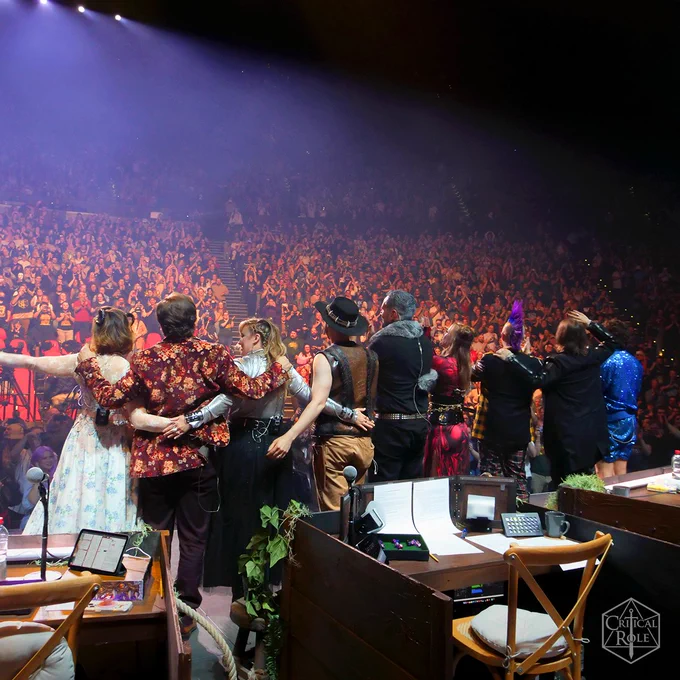 The cast of Critical Role pose on stage in front of a huge crowd of people in a stadium setting in this photo