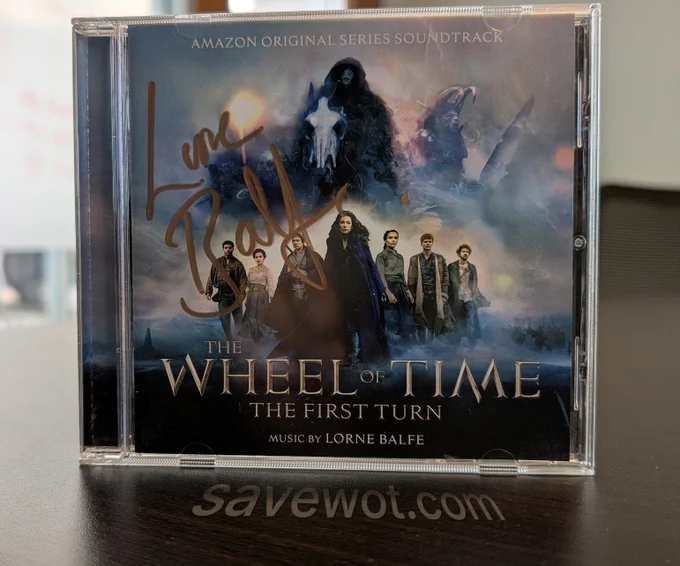 A signed CD copy of The Wheel of Time: The First Turn soundtrack, part of the Amazon Original Series Soundtrack. The cover features main characters from the show, including Moiraine, Lan, Egwene, Rand, Perrin, Nynaeve and Mat, set against a mystical, blue-toned backdrop with shadowy figures. The cover is autographed in gold marker by composer Lorne Balfe. Savewot.com was photoshopped to appear on the table in front of the CD
