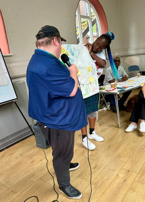 Image of a man and woman standing next to a piece of flip chart paper that has drawings a sticky notes on it. The man is speaking into a microphone. 