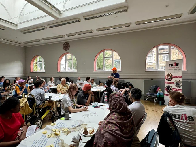 Large meeting hall filled with people sitting around rectangular tables. One person is standing at a table and speaking into a microphone while everyone listens.