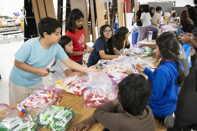 Children sorting and organizing colorful markers into plastic bags around a large table in a classroom setting.