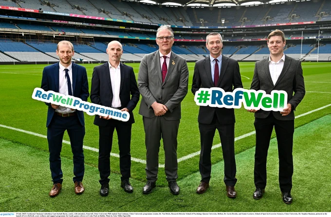 Five men in suits standing on a sports field holding signs promoting the RefWell Programme and #RefWell initiative.