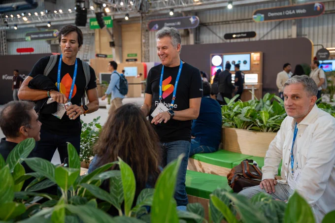 Three people wearing "Google I/O" lanyards and two with "Firebase" t-shirts interact at a tech conference. A person with graying hair and a beard, wearing a white shirt and a "Google I/O" lanyard (name tag "Brandon Badger"), sits smiling on the right. In the center, a smiling person with graying hair and a "Firebase" t-shirt (name tag "Angel Cabrera") engages in conversation. To the left, a person with dark hair and a backpack, wearing a "Firebase" t-shirt (name tag "Seba Gniegrella"), opens a can. Green plants frame the foreground, and event signs like "Demos | Q&A Web" and "Google Pixel" are in the background.