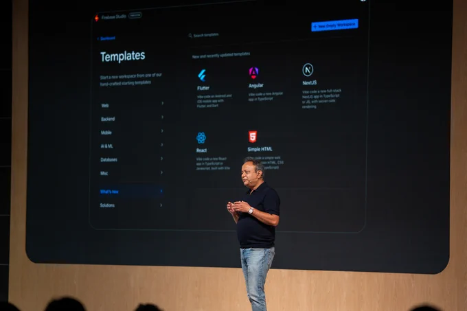A presenter with short dark hair, wearing a dark polo shirt, blue jeans, and a gold watch, stands on a stage with a subtle ear-mounted microphone, gesturing while appearing engaged. Behind them, a large screen displays the "Firebase Studio Templates" user interface, featuring a "New Empty Workspace" button and a list of "New and recently updated templates" for development frameworks such as Flutter, Angular, NextJS, React, and Simple HTML, alongside categories like "Web" and "Mobile."