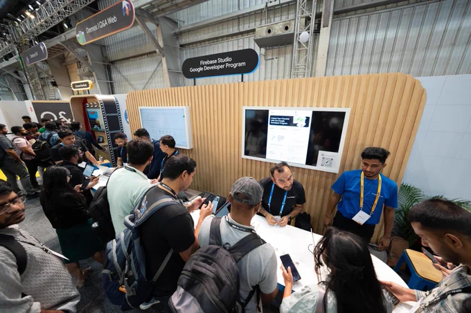 A bustling event booth showcases the "Firebase Studio Google Developer Program" within a large exhibition hall. Numerous attendees, mostly young adults with short, dark hair and wearing casual clothing, gather around white tables, engaging with staff members wearing Google lanyards. One staff member, with a beard and round, rimless glasses, is seated at the table talking to attendees, while another staff member in a blue t-shirt stands to the right. A large screen prominently displays "Unlock Your Potential with the Google Developer Program" along with a QR code to "Join the Google Developer Program," while overhead signs direct visitors to "Demos | Q&A Web" and "Demos | AI."